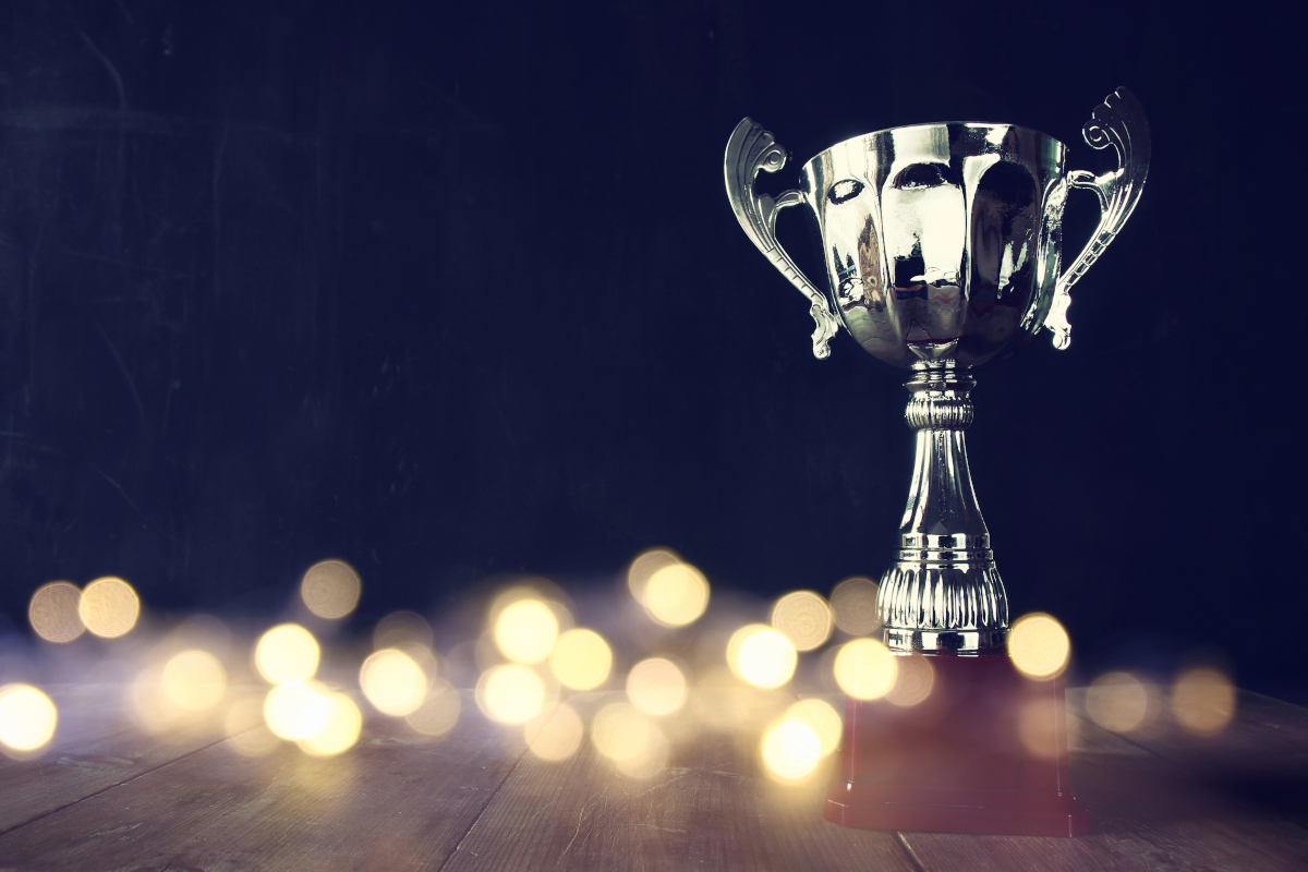 A silver trophy with a wooden base sitting on a wooden-plank surface with out of focus sparks around it.