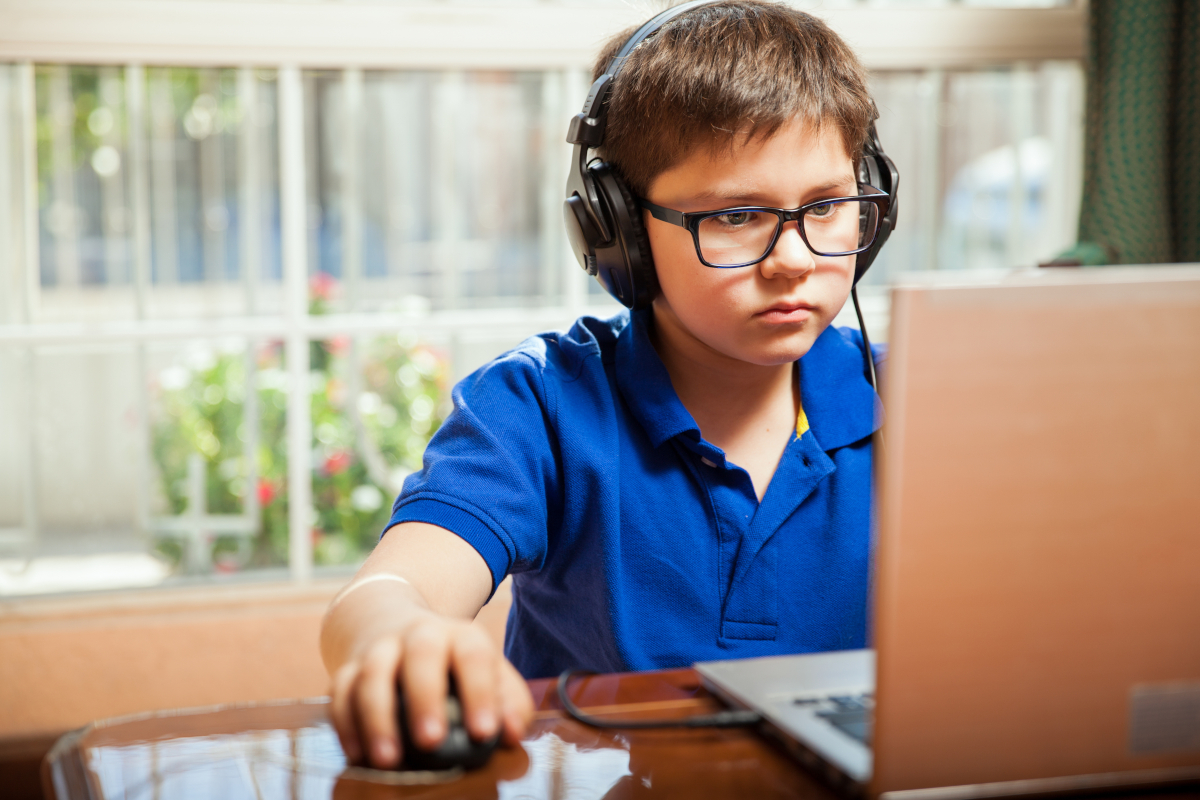A focused child wearing glasses and headphones works on a laptop, using a mouse.