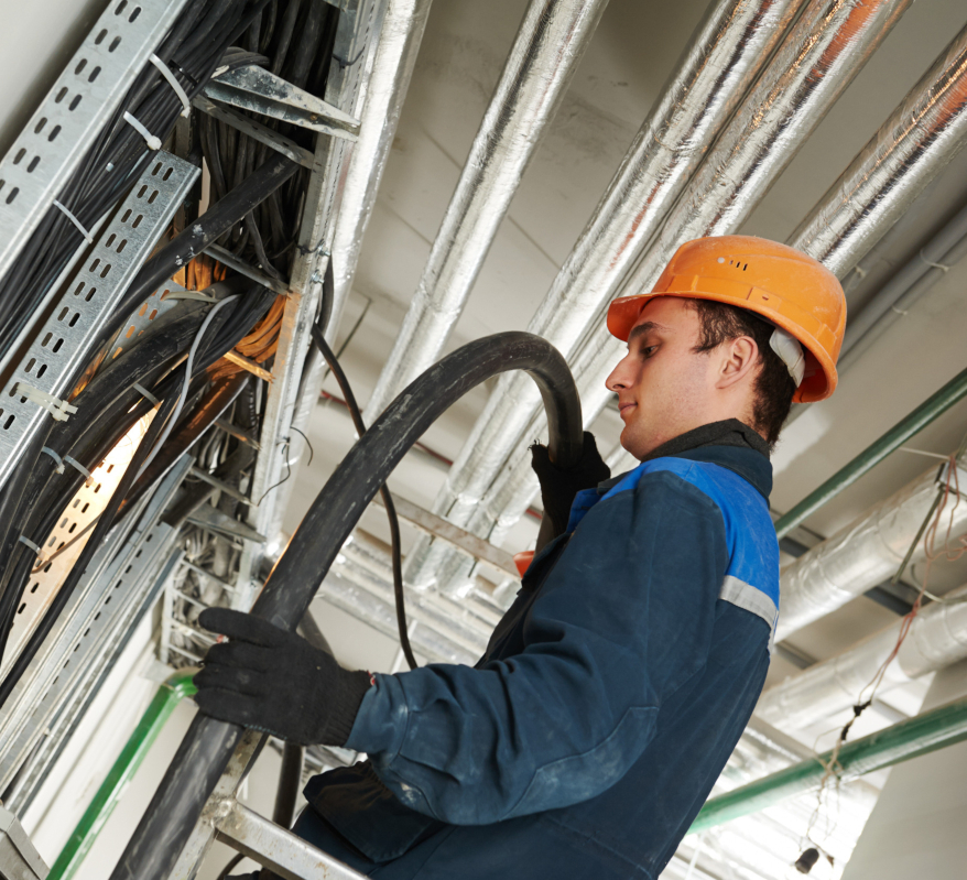 A construction worker in a hard hat and gloves handles a large cable on a ladder. 