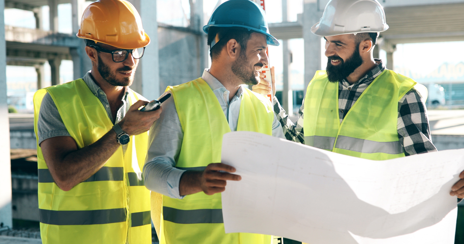 Three construction workers wearing safety vests and helmets discuss a blueprint on a sunny site.
