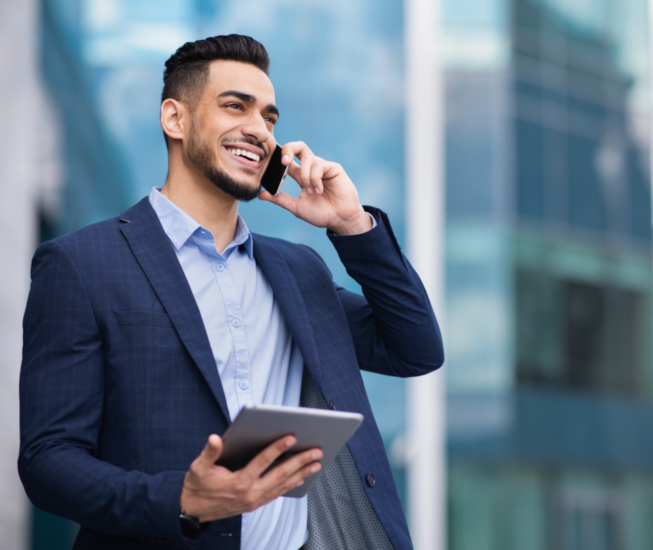 Man in a blue suit smiles while talking on a smartphone and holding a tablet.