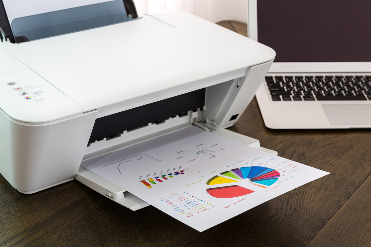A white printer on a wooden desk outputs a colorful chart with graphs and a pie chart, next to a sleek silver laptop.
