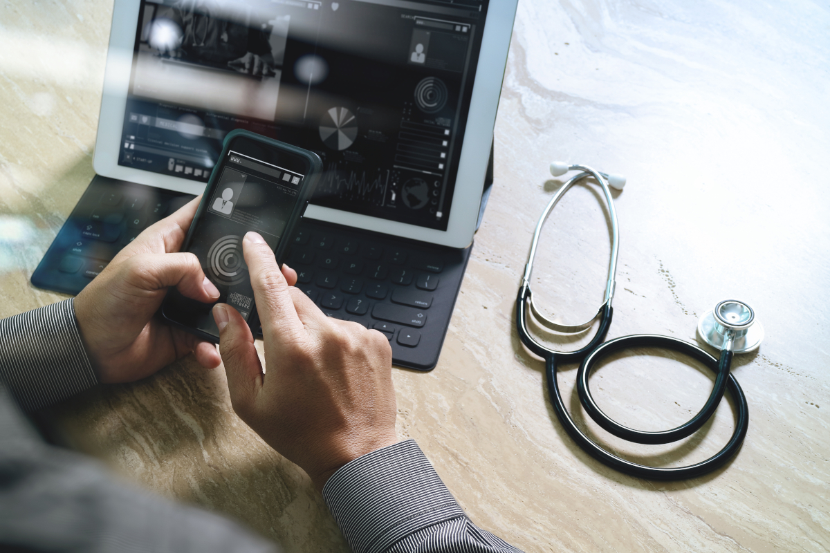 A person's hands hold a smartphone displaying data, with a tablet showing medical info in the background and a stethoscope nearby.