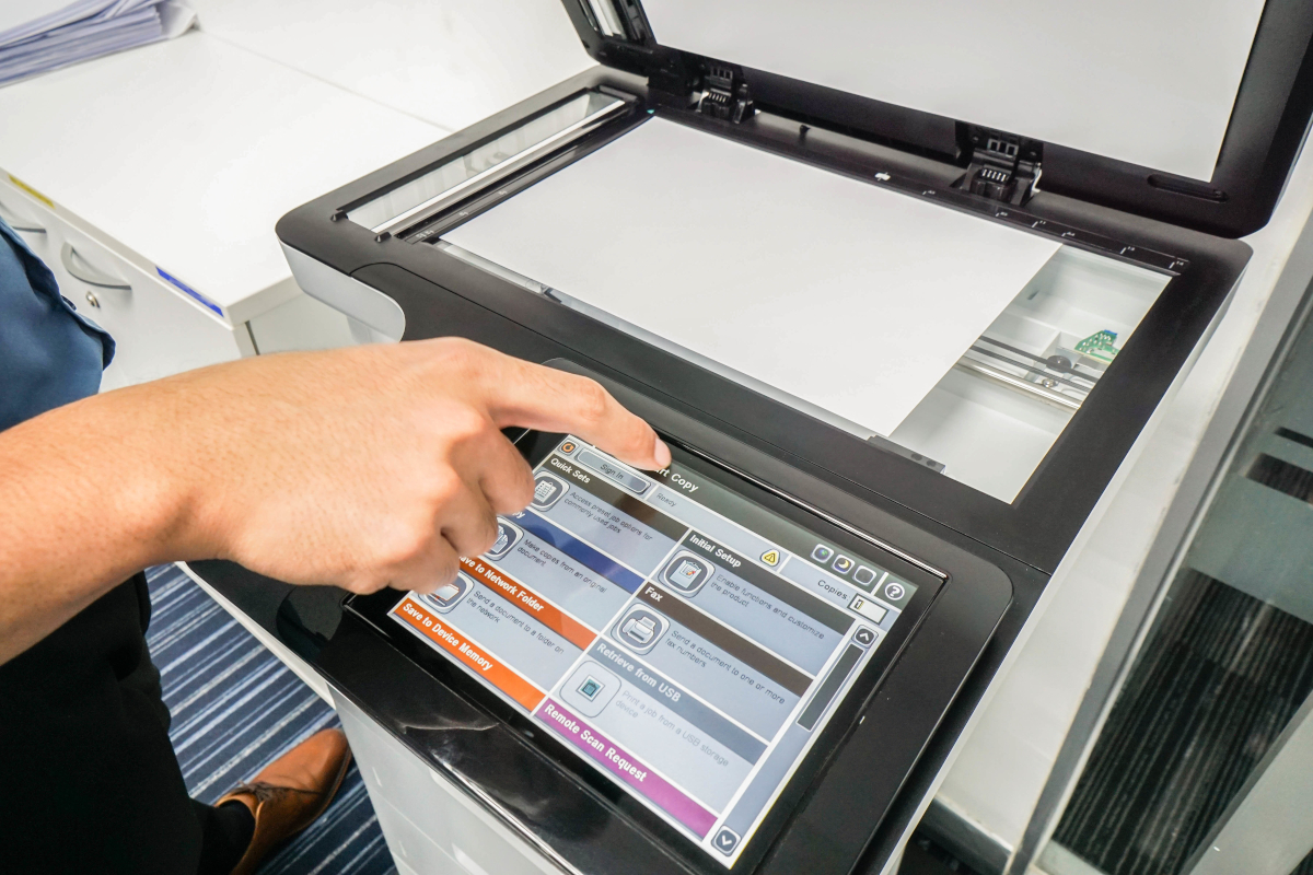 A hand operates a touchscreen interface on a photocopier or scanner. The machine's lid is open, revealing a white document inside. 