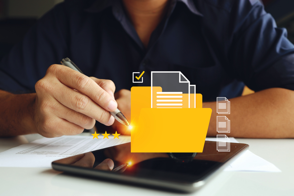 A person in a dark shirt writes with a pen on paper. A digital overlay shows a yellow folder with documents, a checkmark, and stars above a tablet.