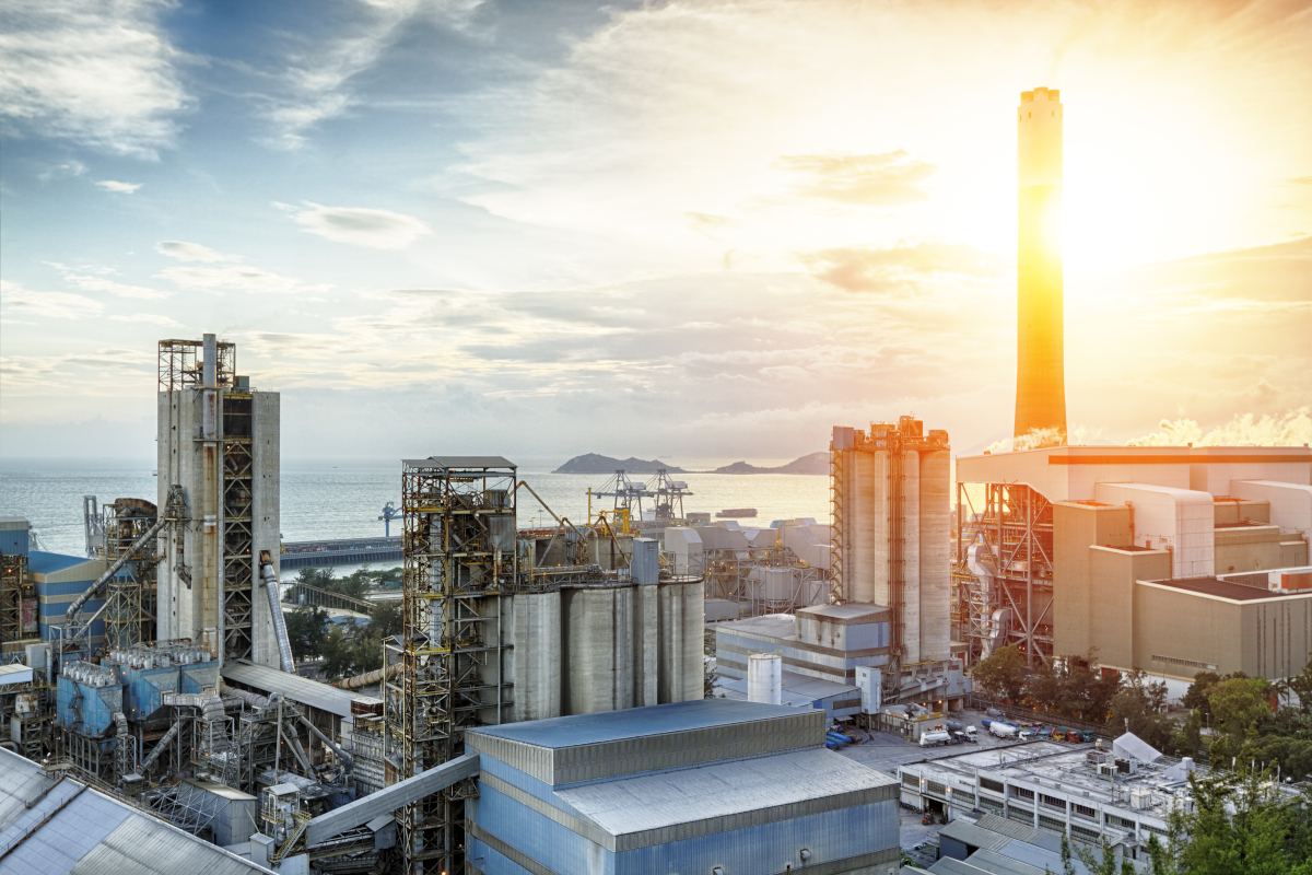 Industrial complex by the sea at sunset, featuring large chimneys and metal structures.