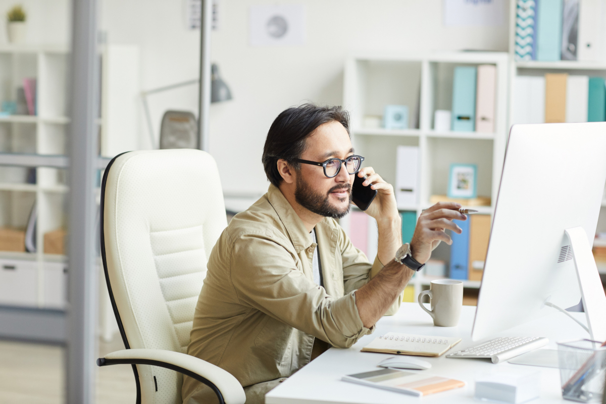 Man with glasses at a desk in a bright office, talking on the phone and using a computer.